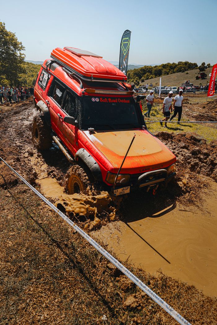 Red 4x4 SUV navigating through muddy terrain during an off-road event with spectators.